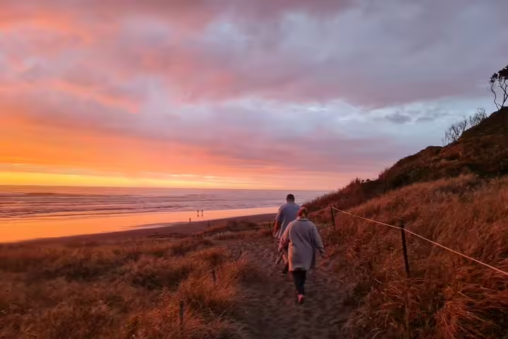 Two people walking along a scenic Auckland beach path during a vibrant sunset, ideal for a romantic tour experience.