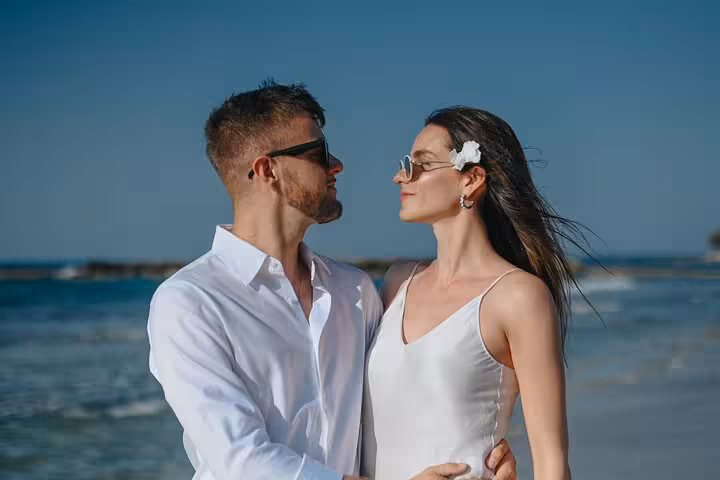 Romantic couple in white attire embracing on Anisaras beach during private photoshoot, with serene ocean backdrop.