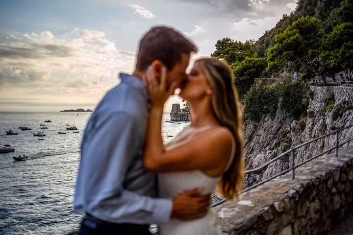 Romantic couple kissing on a scenic cliffside with boats dotting the sea during a sunset Amalfi Coast photo shoot.