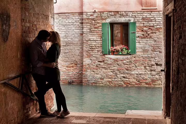 Romantic couple kissing in a quaint Venetian alley with a picturesque canal and flowered window in the background.