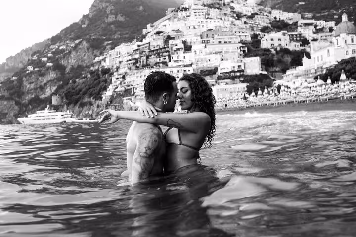 Romantic couple embracing in the sea with Positano's iconic hillside and a yacht in the background, ideal for Amalfi memories.