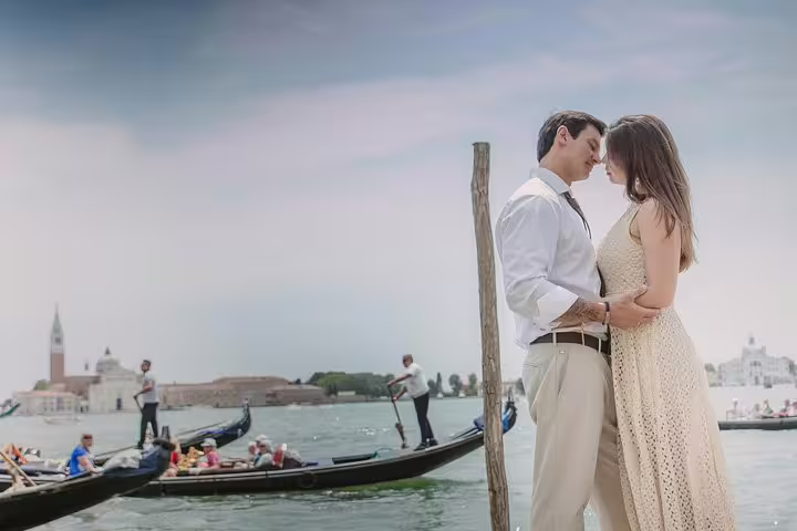 Romantic couple embracing near gondolas on a serene Venetian canal with historic architecture in the background.