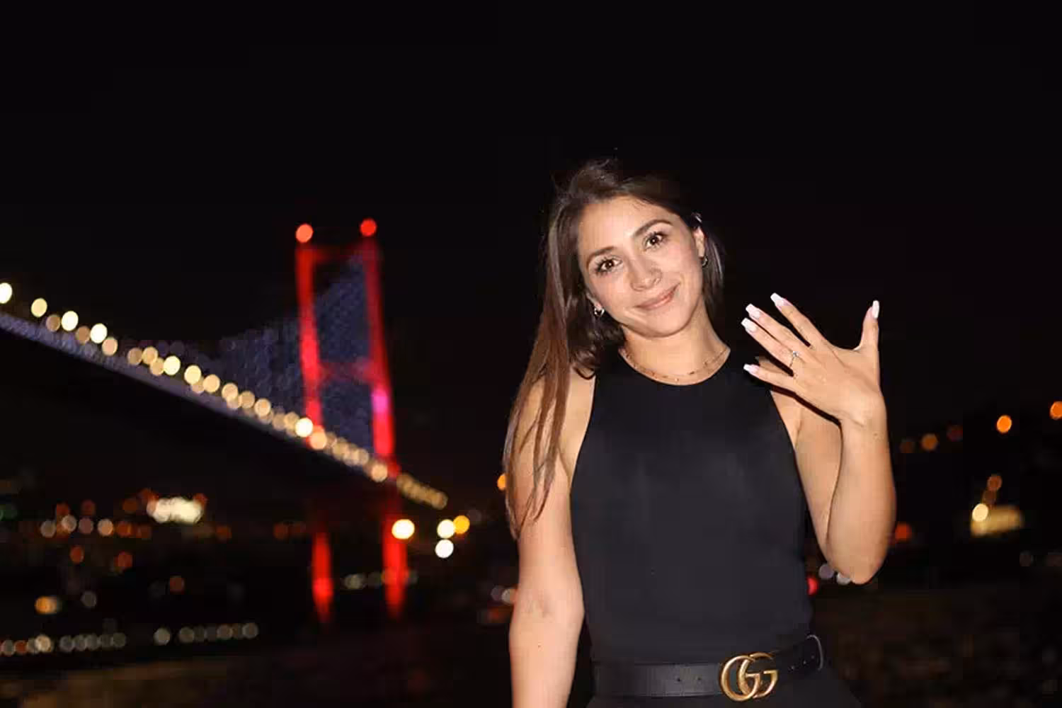 Woman showing engagement ring during romantic marriage proposal experience with illuminated bridge at night backdrop