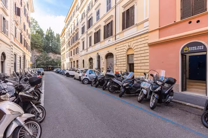 Urban street in Rome featuring Rome Bag Storage entrance with scooters and cars parked nearby.