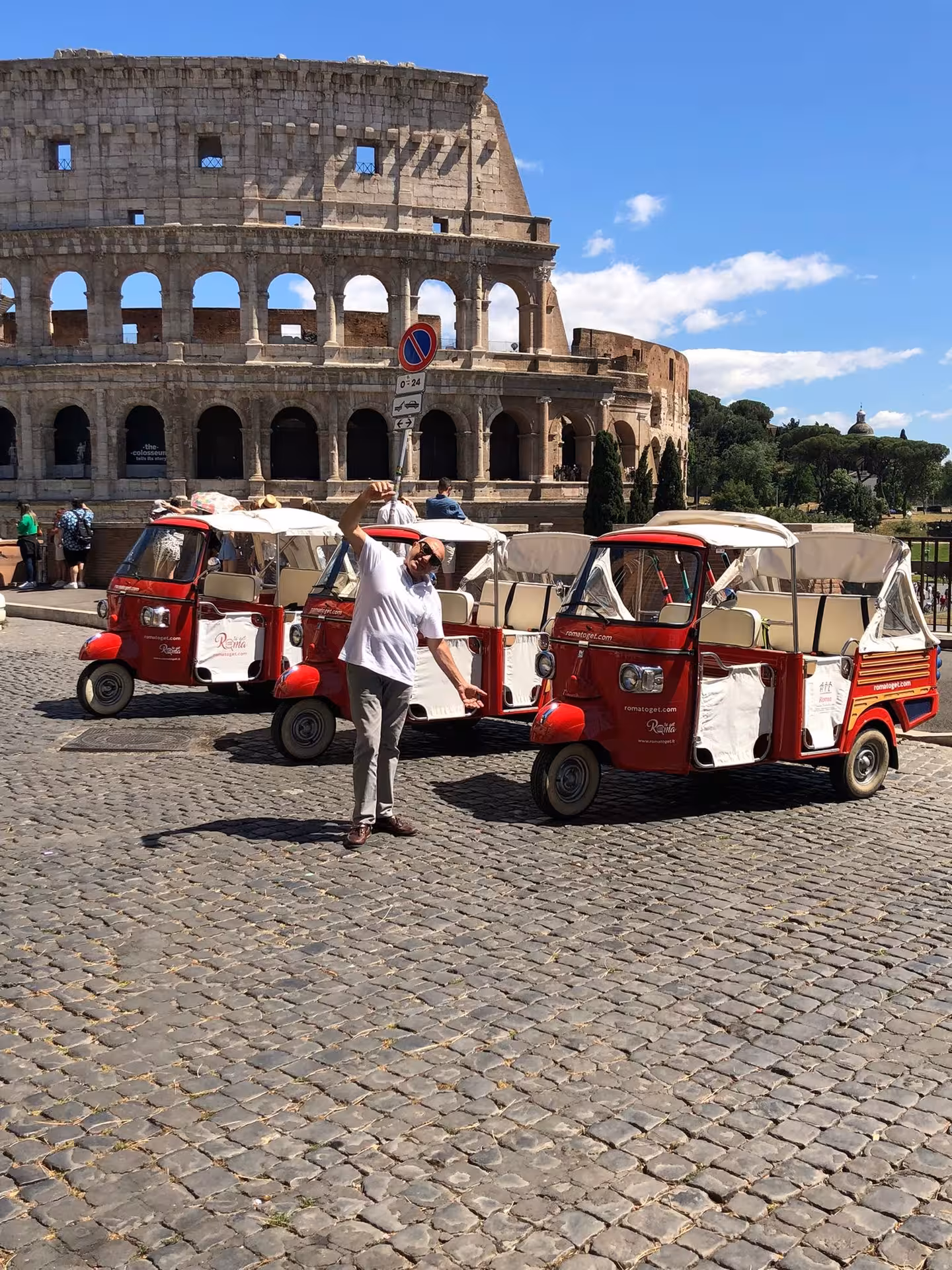 Tourists pose with red tuk tuks in front of the iconic Colosseum in Rome under a bright daytime sky.
