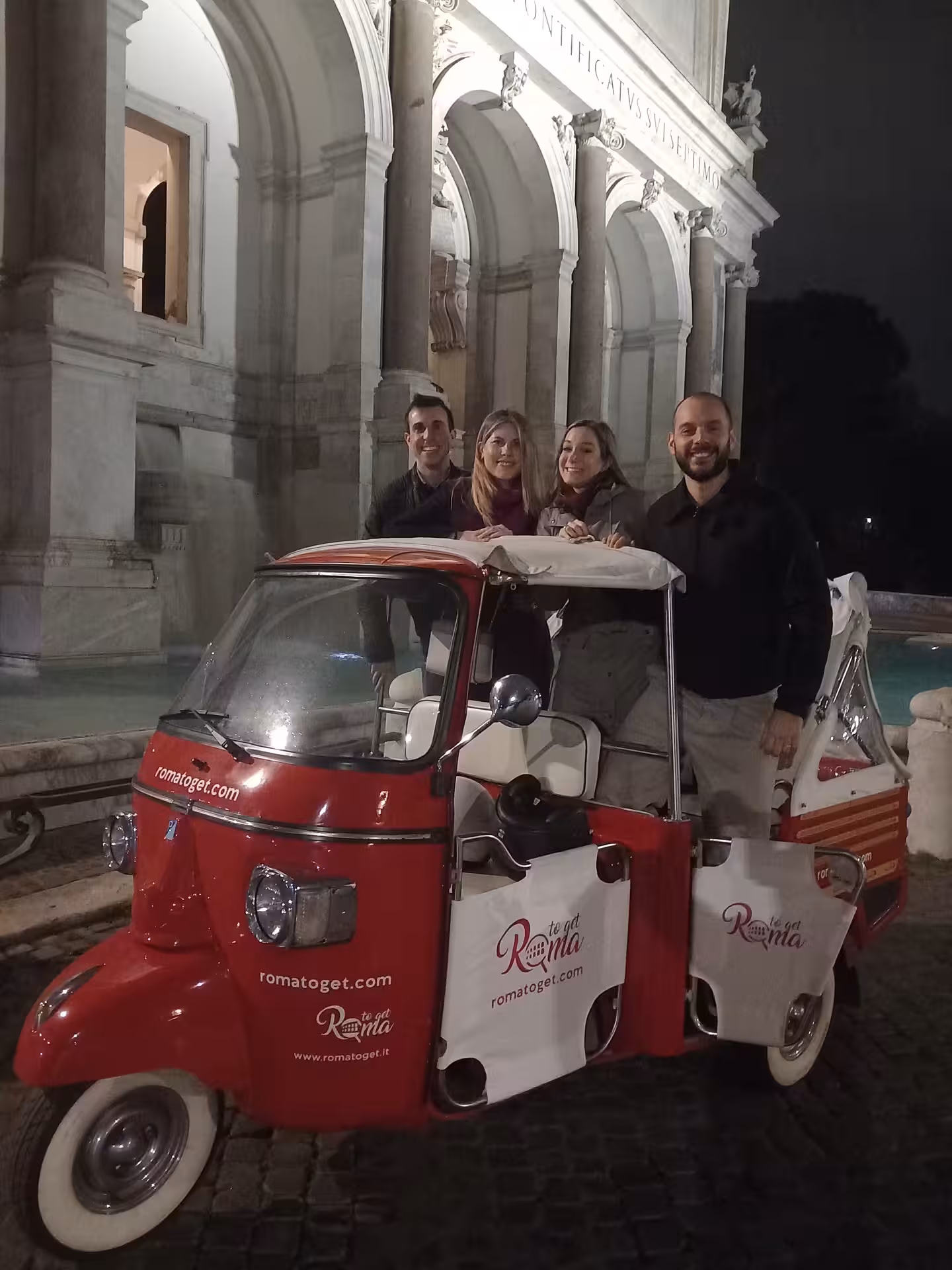 Tourists enjoy Rome by night in a red tuk tuk near a historic monument, capturing the city's vibrant atmosphere.