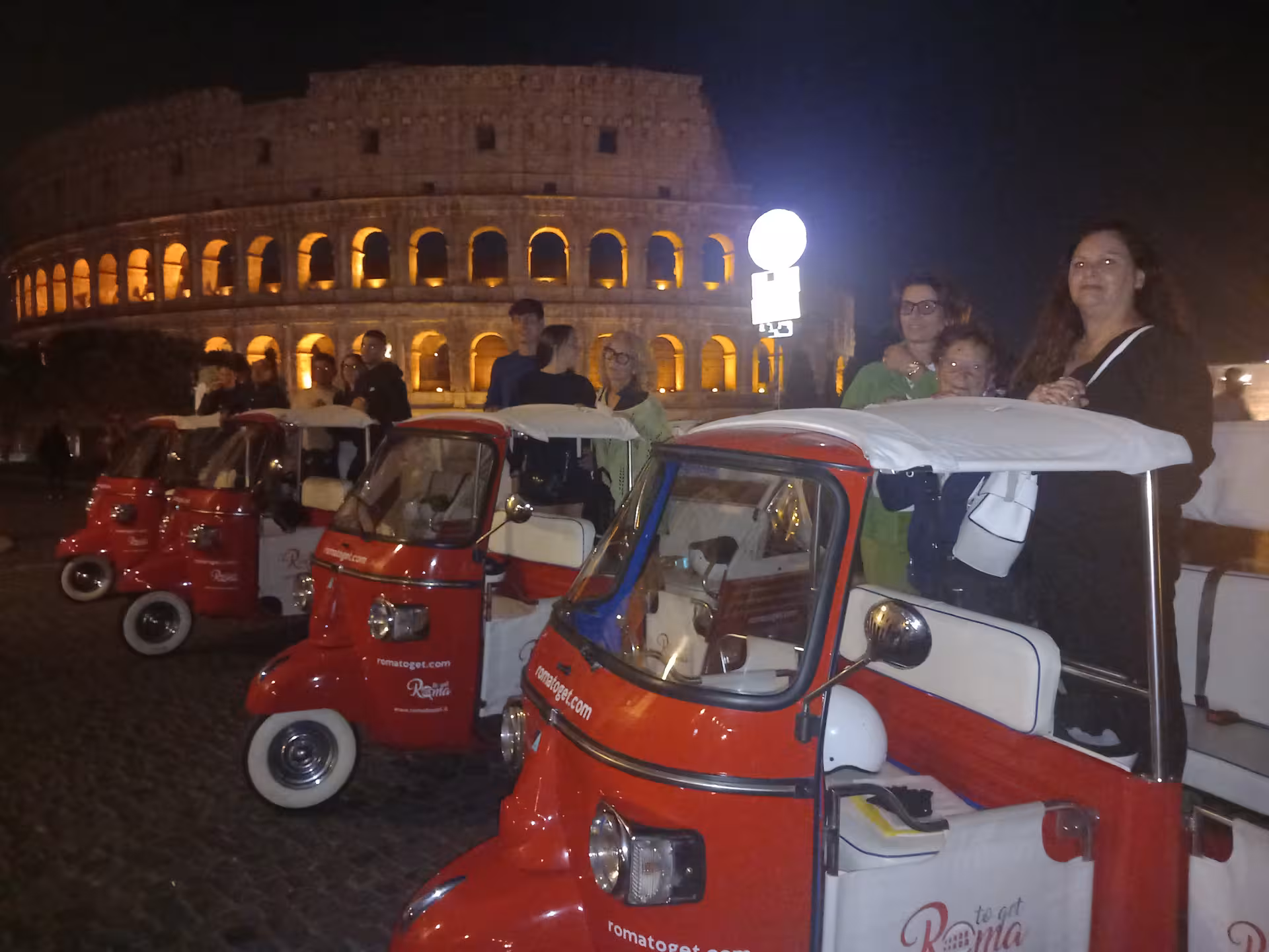 Visitors enjoy a night tour with illuminated red tuk tuks at the Colosseum, Rome's historic landmark.