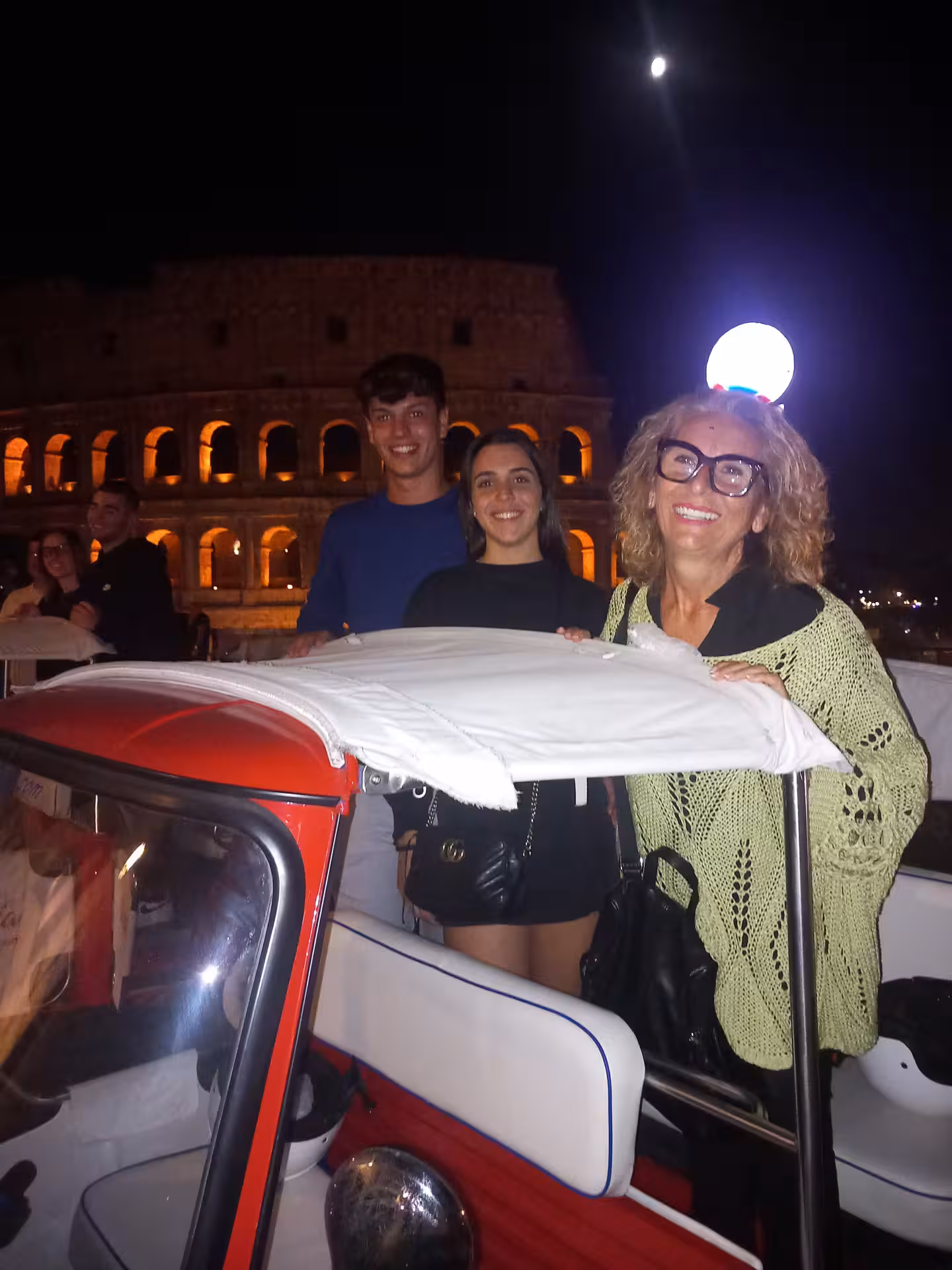 Smiling group enjoying a night tuk tuk ride with the Colosseum glowing in the background under the moonlight.