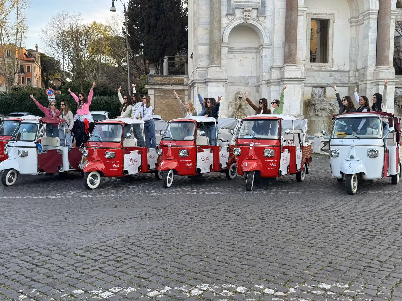 Group enjoying a Rome tour with multiple colorful Piaggio ape calessinos parked in front of a historic monument.