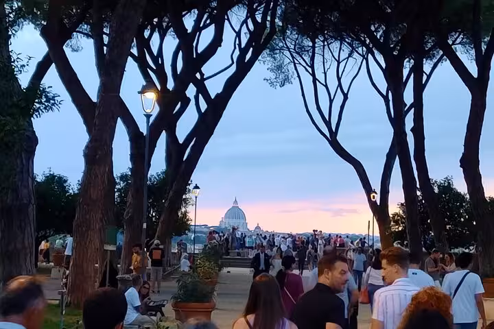 Tourists admire the sunset view of St. Peter's Basilica from a tree-lined path in Rome.