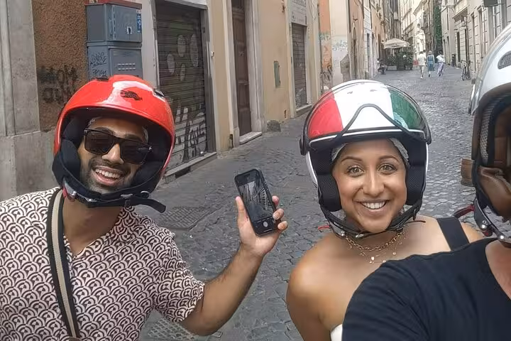 Smiling tourists wearing helmets capture a selfie during a guided Vespa tour through the charming streets of Rome.