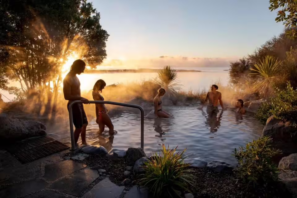 Tourists relaxing in a geothermal hot spring at sunset in Rotorua, part of the Auckland luxury tour.