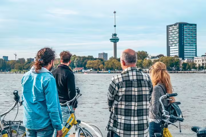Rotterdam bike tour guests pause by the Maas River, viewing the Euromast and skyline with a local guide