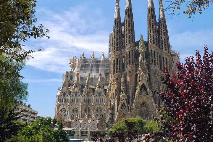 View of the iconic Sagrada Familia's intricate facade surrounded by lush greenery under a clear Barcelona sky.