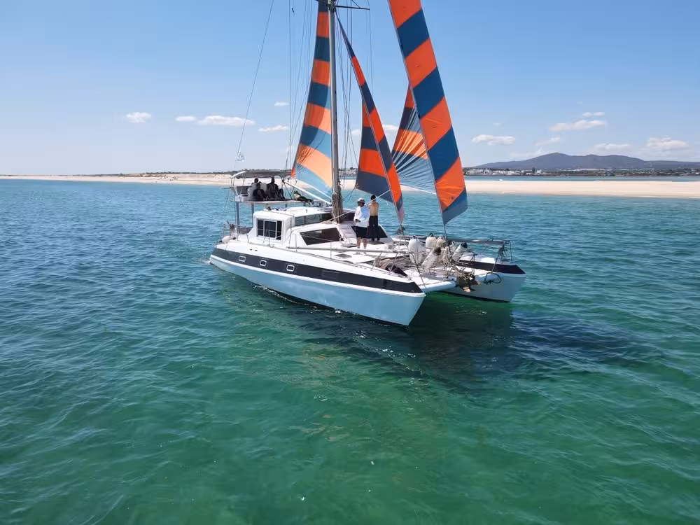 Sail catamaran cruising turquoise Blue Lagoon waters on a half-day tour, with sandy beach and colorful sails