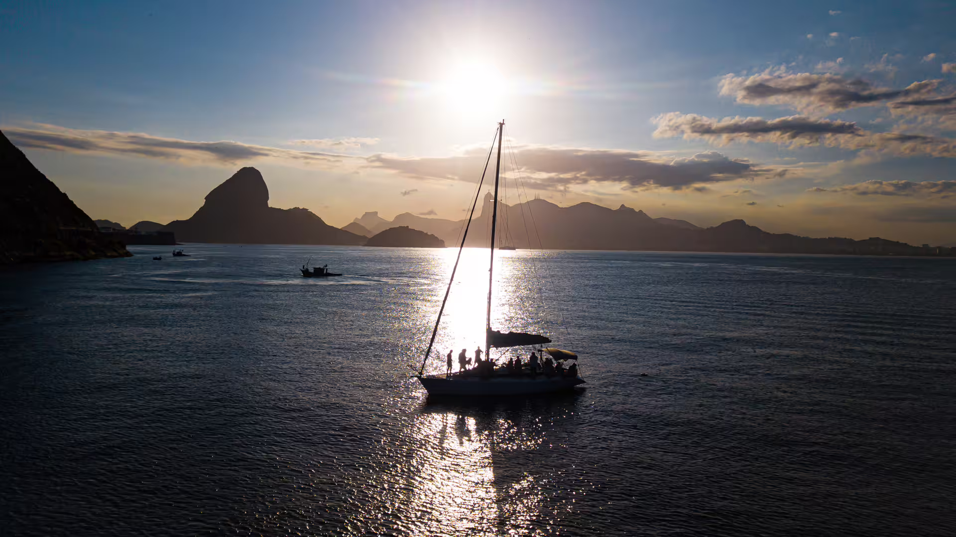 Silhouetted sailboat with passengers at sunset in Cagarras Archipelago, offering a picturesque view of distant mountains.