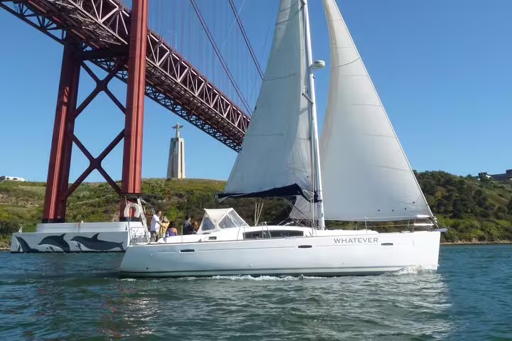 White sailboat cruising under the 25 de Abril Bridge on the Tagus River near Lisbon's famous landmarks.