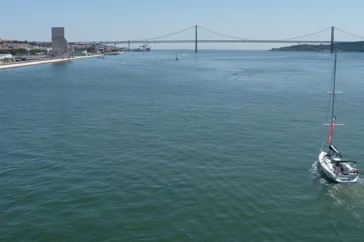 Sailboat gliding on the Tagus River with Lisbon's iconic suspension bridge in the background under clear skies.