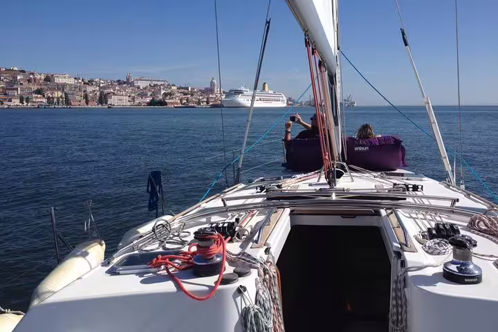 View from a sailboat deck on the Tagus River with Lisbon's cityscape and a cruise ship in the distance.