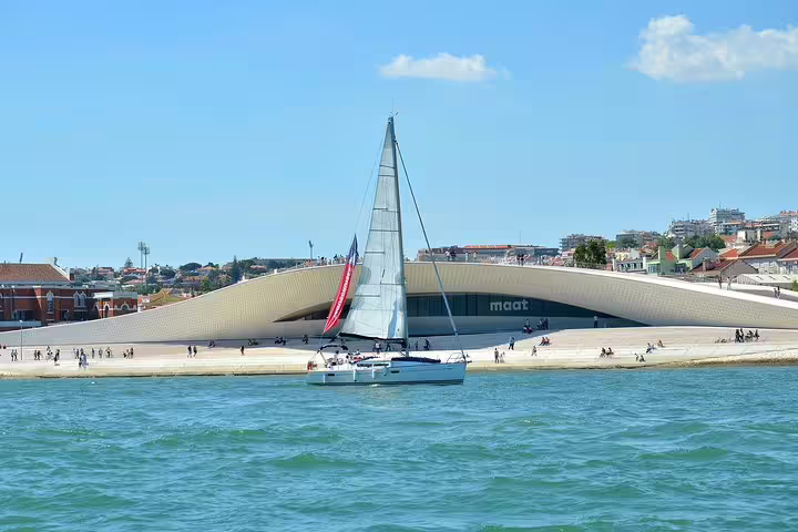 Sailboat gliding past the MAAT museum on the Tagus River with a backdrop of Lisbon cityscape and blue skies.