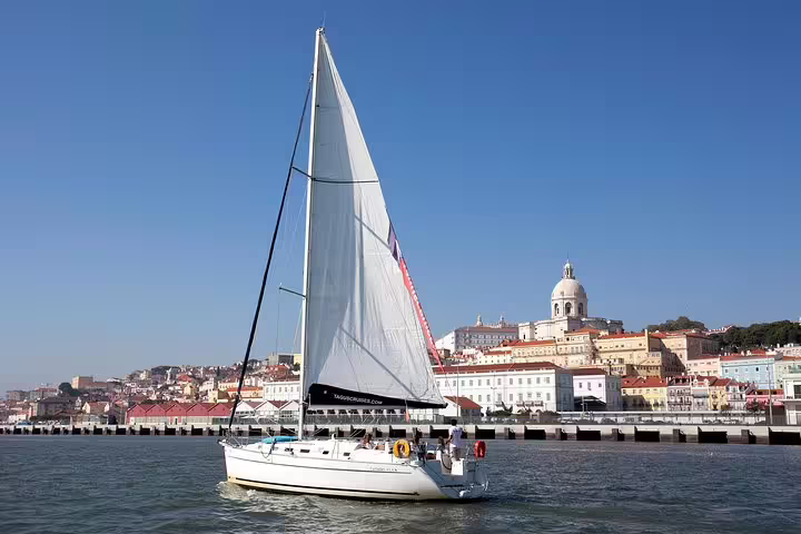 Sailboat gliding on the Tagus River with Lisbon's historic skyline and dome in the background under a clear blue sky.