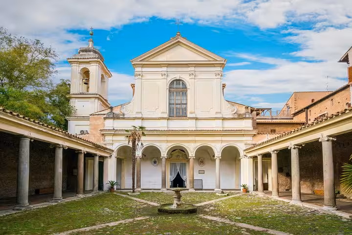 Peaceful cloister courtyard of San Clemente Basilica in Rome, with central fountain, columns, and historic church facade under blue sky