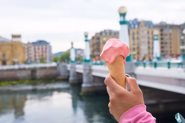 Hand holding a pink ice cream cone with San Sebastian's picturesque cityscape and bridge in the background.