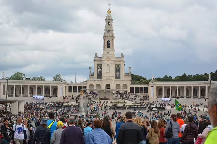 Crowds gather at the Sanctuary of Fatima in Portugal, a key pilgrimage site, with its iconic basilica under a cloudy sky.