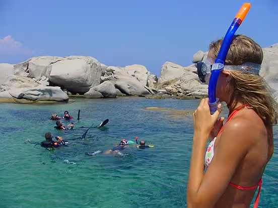 Woman with snorkel gear overlooks group diving in Santa Teresa di Gallura's turquoise waters, surrounded by rocky cliffs.