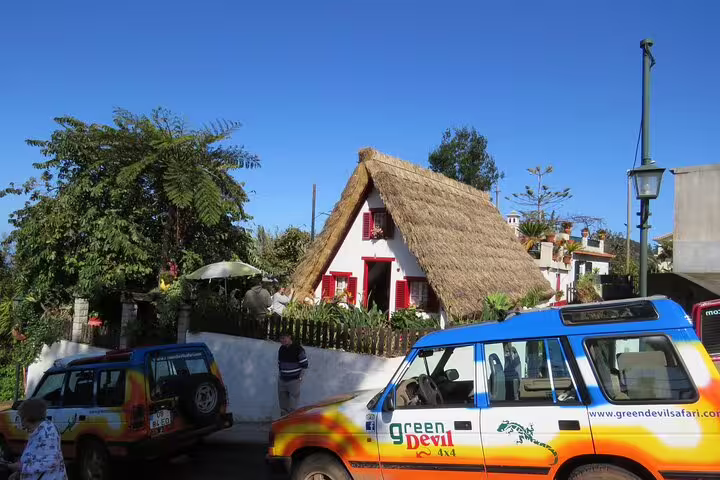 Traditional Santana house with a thatched roof in Madeira's northeast, featuring vibrant tour jeeps under a clear blue sky.