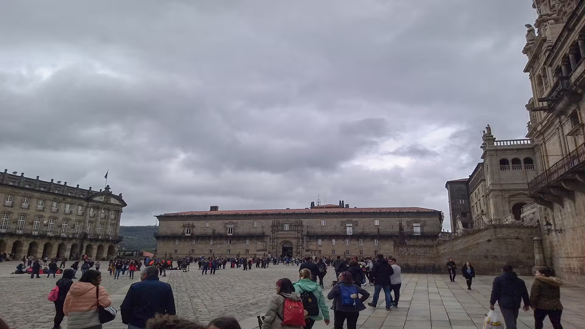Visitors explore the bustling square in front of the historic Santiago de Compostela Cathedral under a dramatic cloudy sky.