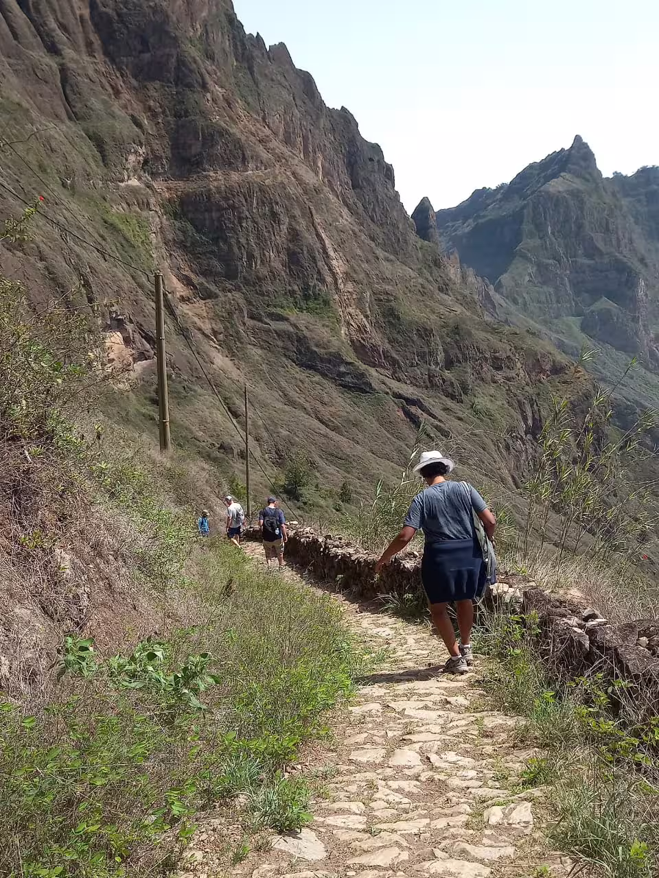 Hikers explore the scenic trails of Santo Antão's Valley of Paul, surrounded by rugged cliffs and lush greenery on a sunny day.