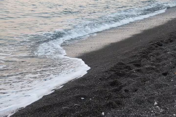 A serene view of Santorini's black sand beach with gentle waves, ideal for a private full-day sightseeing tour experience.