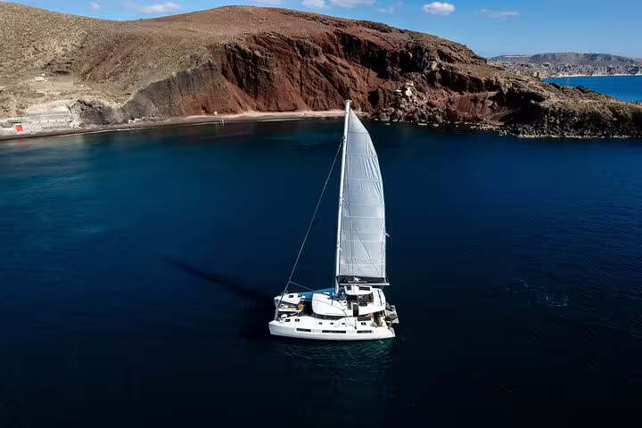 Sailing catamaran gliding through the Aegean Sea near Santorini's iconic red cliffs on a half-day cruise.
