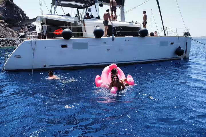 Guests enjoy swimming and relaxing on a flamingo float near a luxurious catamaran on a Santorini cruise.