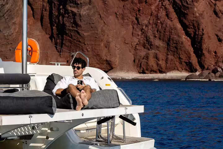 Relaxed man enjoying the sun on a luxurious catamaran cruise with stunning Santorini cliffs in the background.