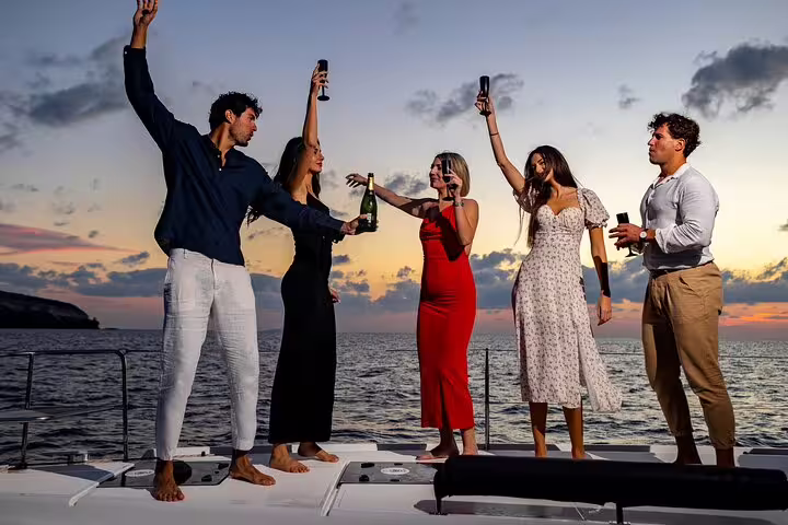 Group of friends enjoying a sunset celebration with drinks on a Santorini catamaran cruise with open bar.