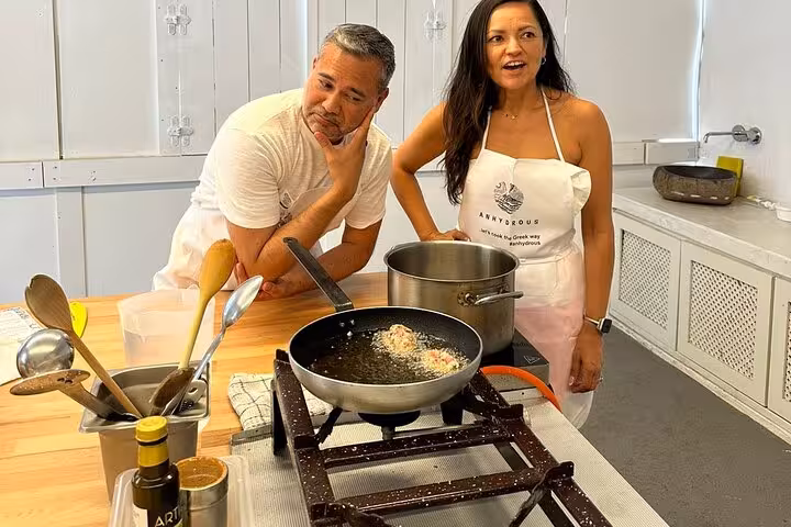 Couple cooking together in Santorini kitchen class, sautéing Greek dish before visiting two local wineries