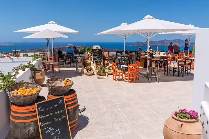 Sunny terrace cafe in Santorini overlooking the Aegean Sea, with vibrant orange chairs and local menu boards, perfect for a relaxing tour stop.