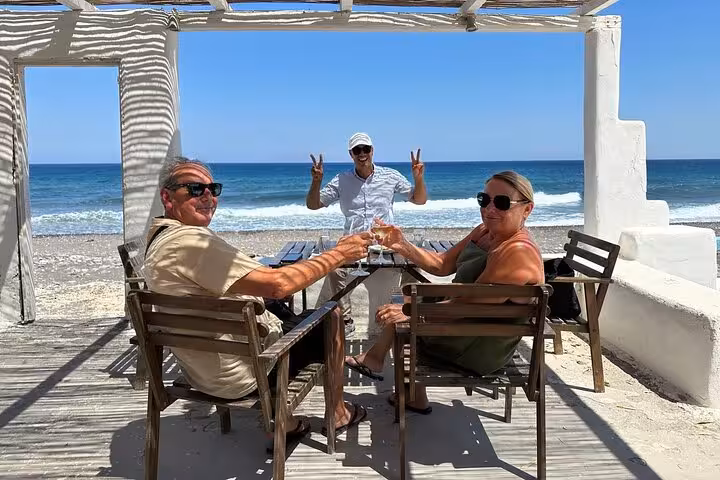 Guests toast seaside at Santorini beach taverna during Island of Thera 8h tour with lunch and wine tasting