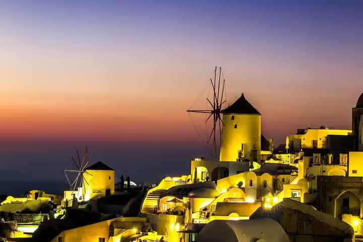 Santorini's iconic windmills and illuminated whitewashed buildings glow under a vibrant sunset sky during a private night tour.