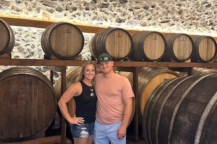 Guests pose by oak wine barrels in a Santorini winery cellar on the 2-winery sunset wine and tapas tour