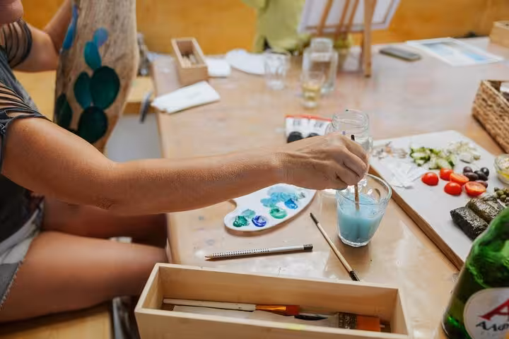 Hands mixing paint beside Greek wine and mezze platter at Santorini wine and paint experience, art supplies on table