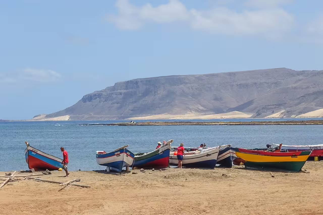 Colorful fishing boats on a sandy beach with people nearby, set against the rugged coastal landscape of São Vicente, Cape Verde.