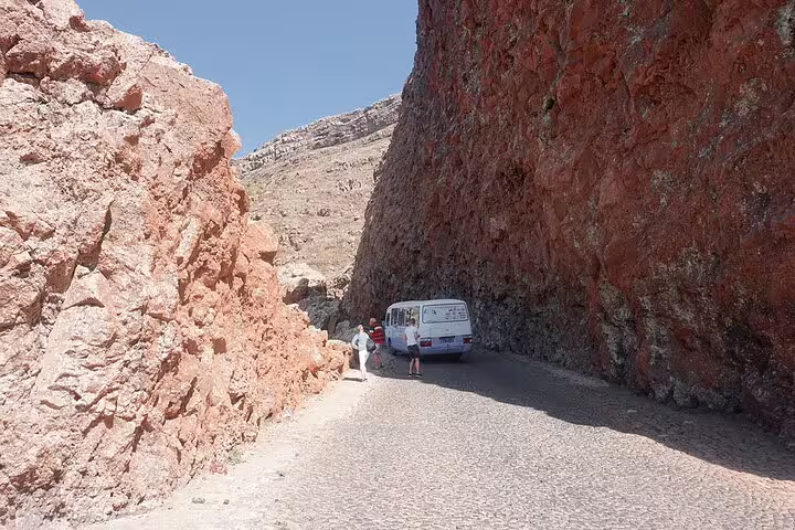 Tourists explore a narrow canyon road on São Vicente Island, surrounded by towering red cliffs under a clear blue sky.
