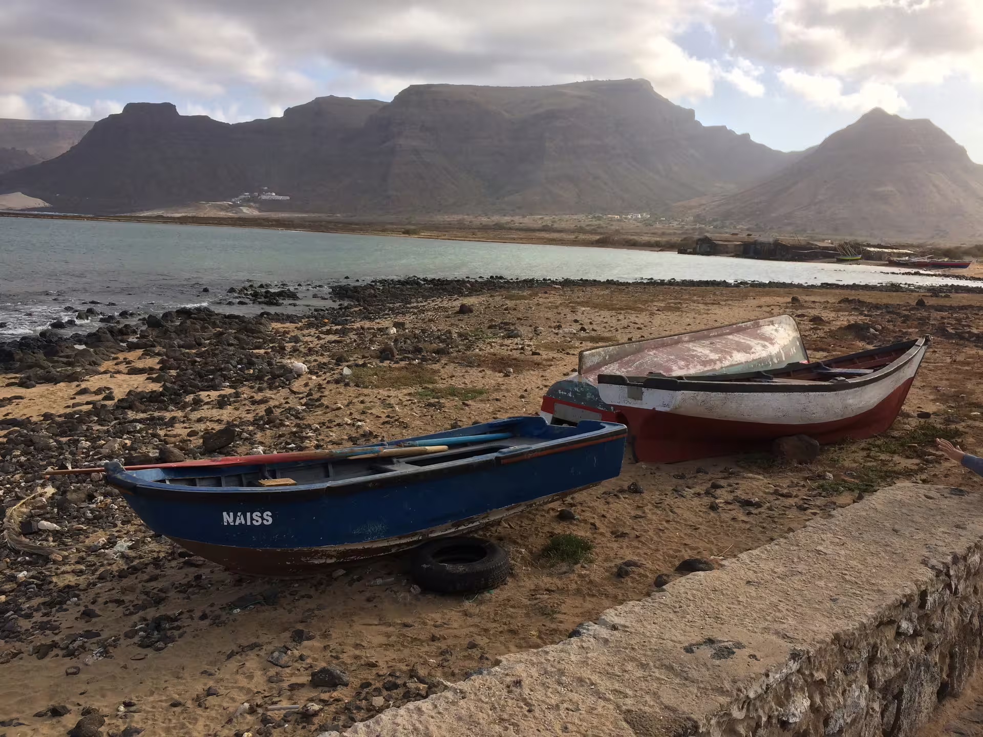Rustic fishing boats rest on a rocky São Vicente beach with majestic mountains in the background, perfect for a Mindelo tour.