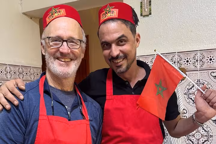 Two participants in red aprons and Moroccan hats enjoying Sardin Tawa Cooking Class Marrakesh, holding a Moroccan flag.