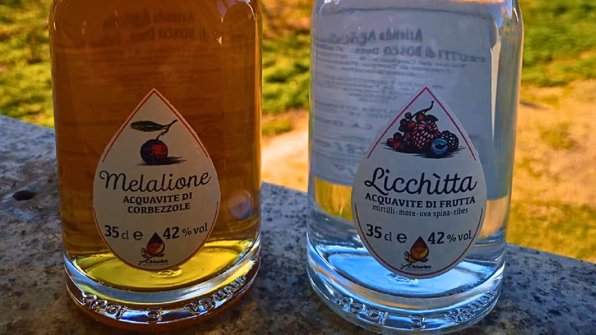 Two bottles of Melalione and Licchìtta fruit distillates on a stone ledge, showcasing Sardinian flavors.