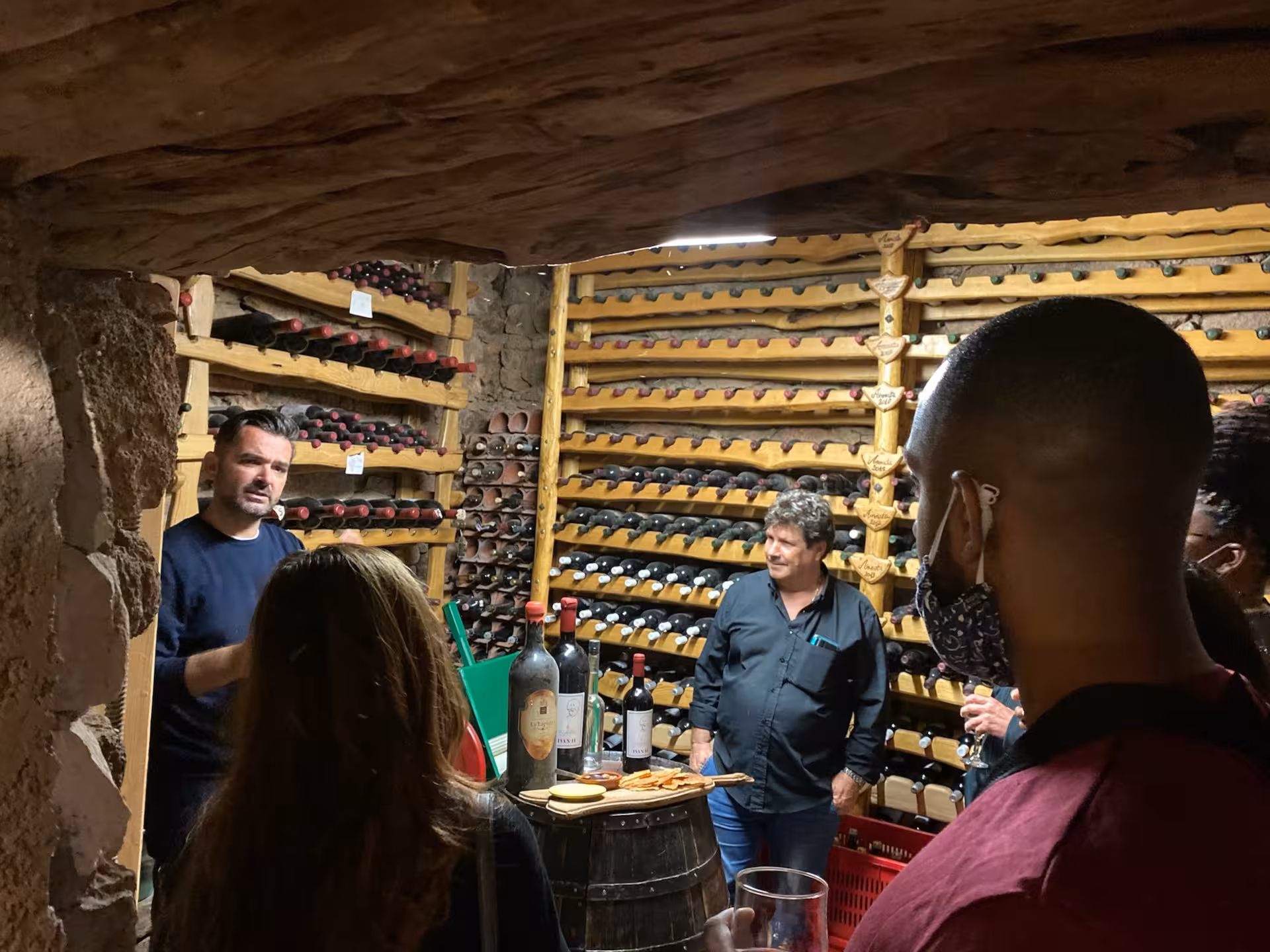 Tour group explores a traditional Sardinian wine cellar lined with bottles during an Olbia pasta workshop.