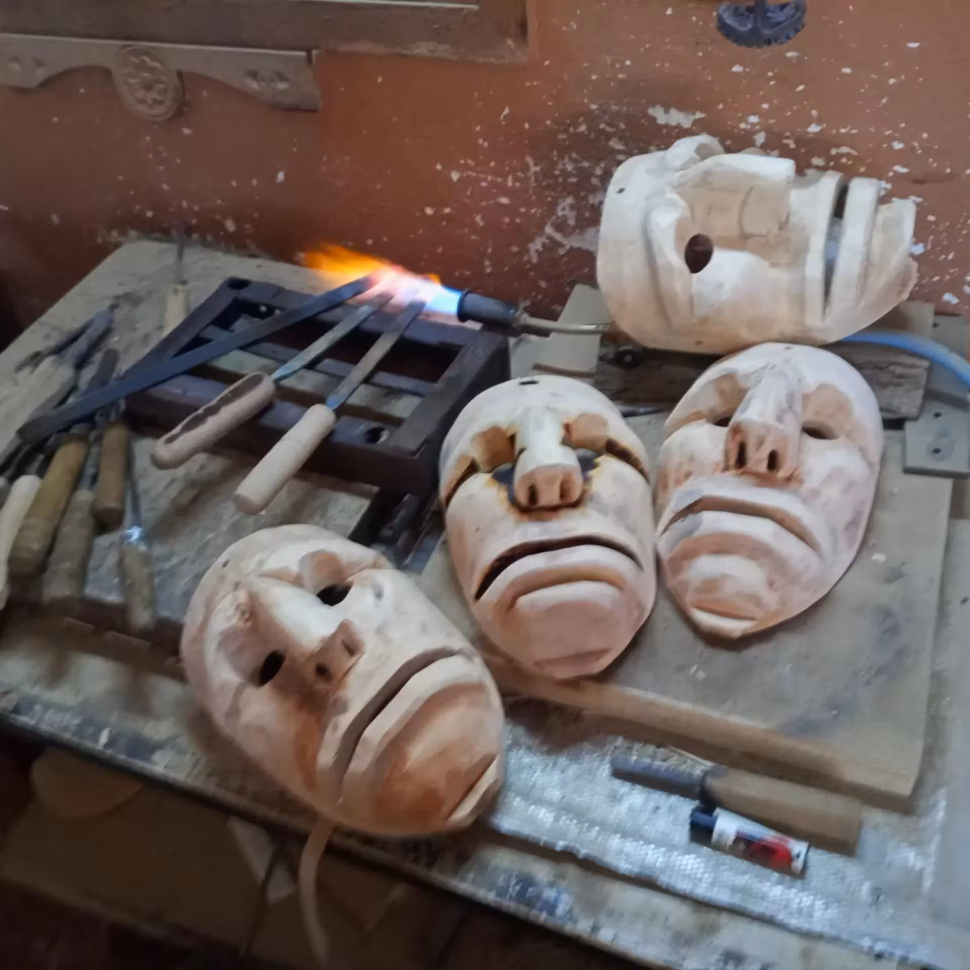 Wooden Sardinian masks being crafted with tools and a gas burner in a Mamoiada workshop.